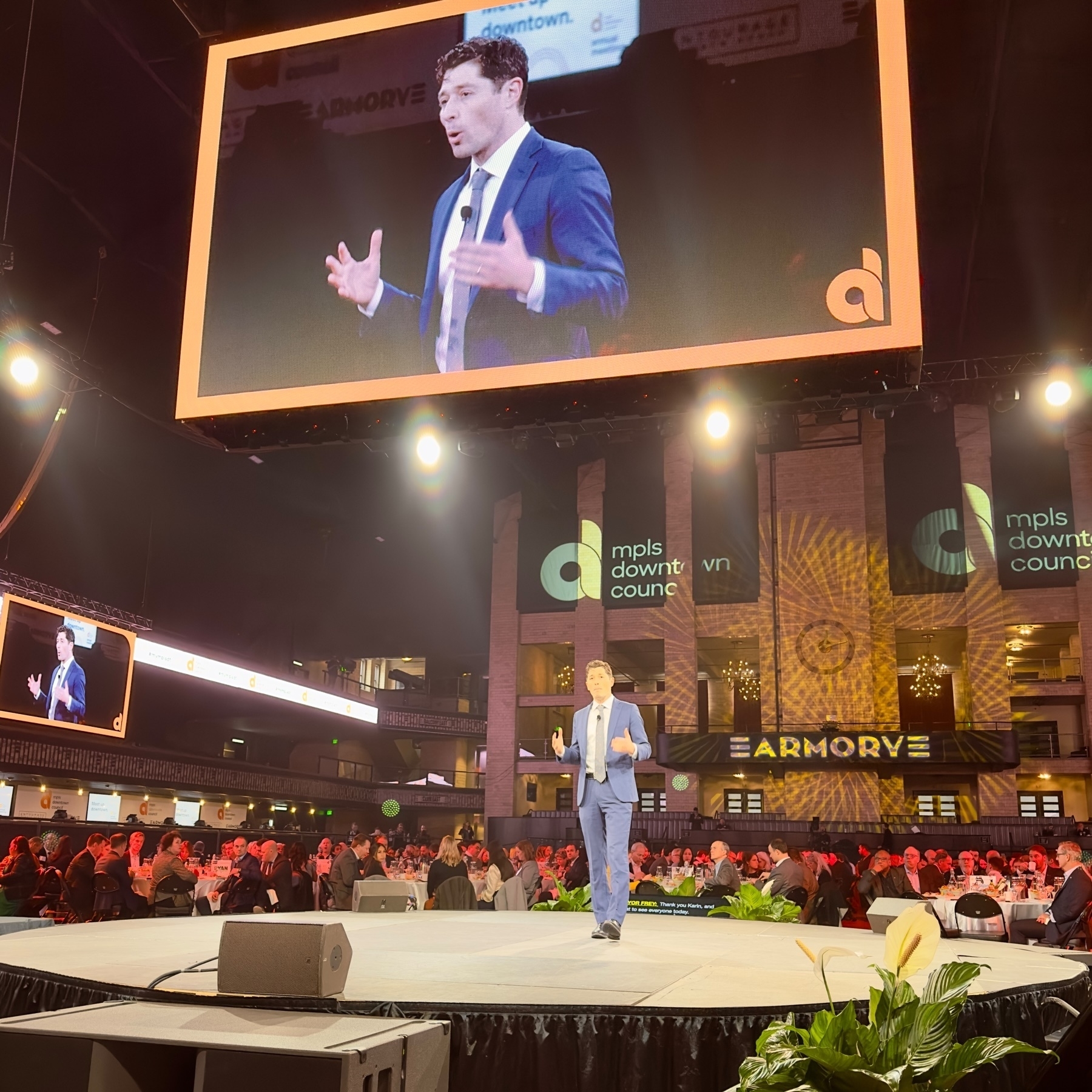 Mayor Jacob Frey on stage at an event, with their image displayed on a large screen behind them. The audience is seated at tables in a large venue, with banners reading “mpls downtown council” and “ARMORY” visible in the background.