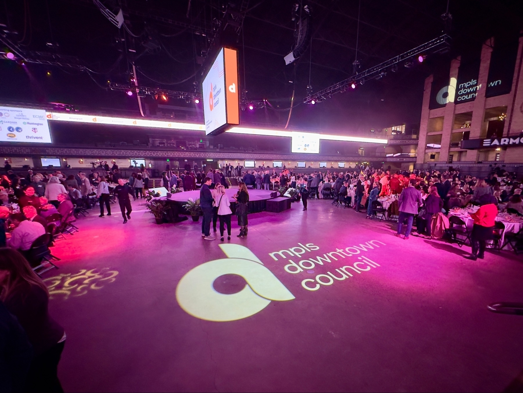 A large indoor event with crowds of people milling around tables, a projection screen displaying information, and the logo “mpls downtown council” illuminated on the floor.