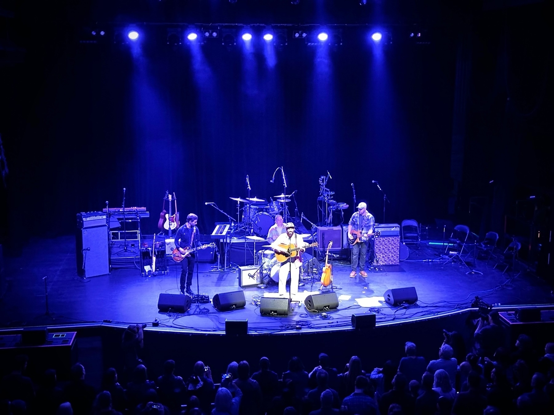 A live concert setting with musicians performing on stage in front of an audience. The stage is lit with blue lighting and features a seated guitarist in the center, with standing band members on either side. There are guitars and other musical equipment arranged around the performers