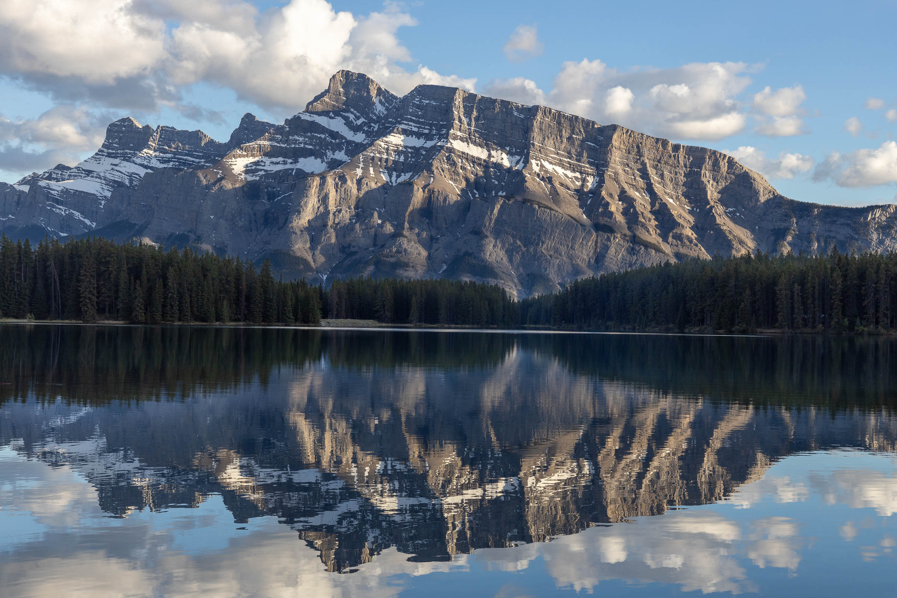 A scenic mountain range is reflected in the still waters of a lake under a sky dotted with clouds.