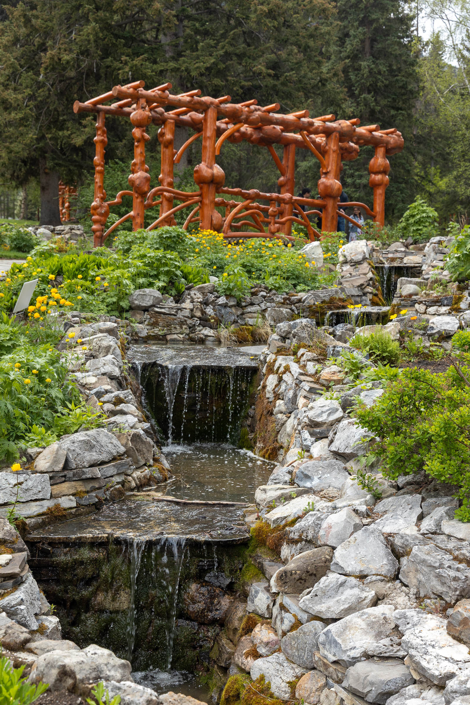 A small waterfall flows through a rocky garden with greenery and a unique wooden sculpture in the background.