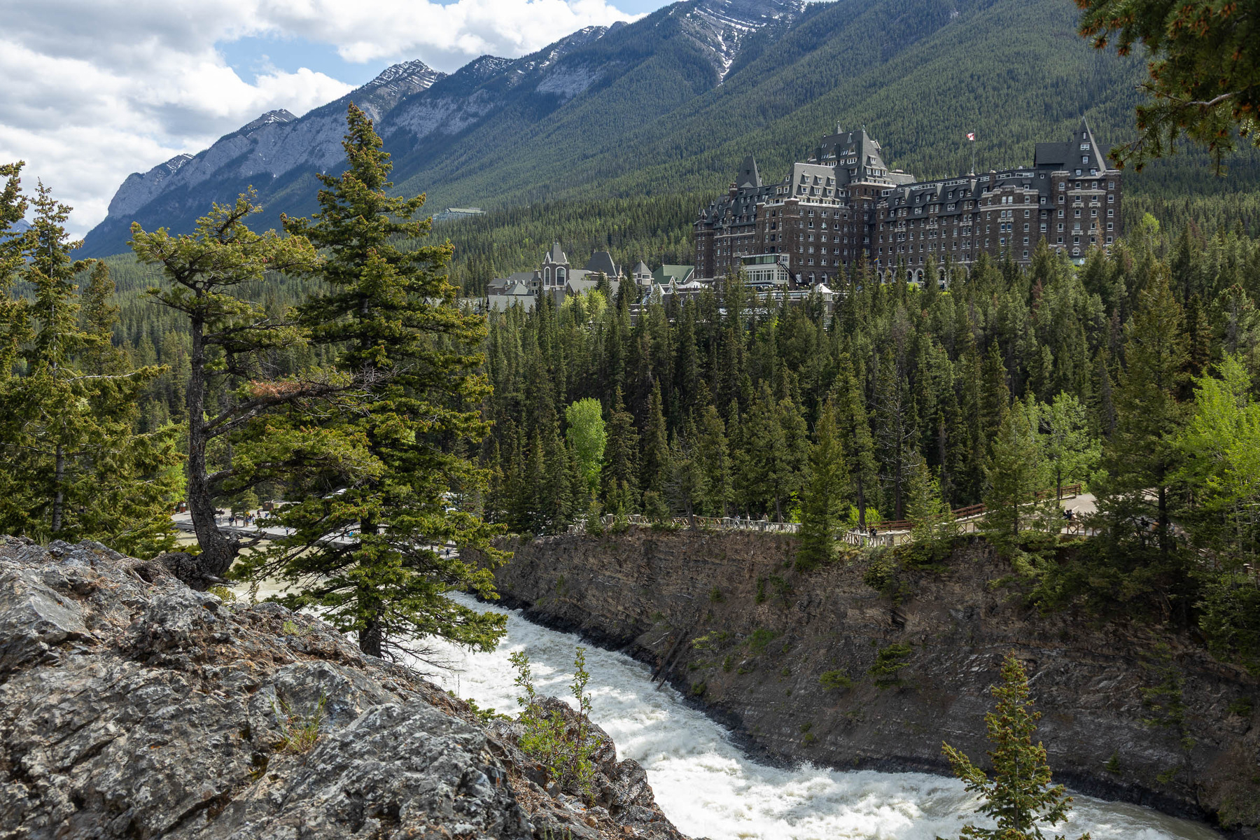 A grand, castle-like hotel is nestled among dense evergreen forests and rugged mountains, overlooking a river.