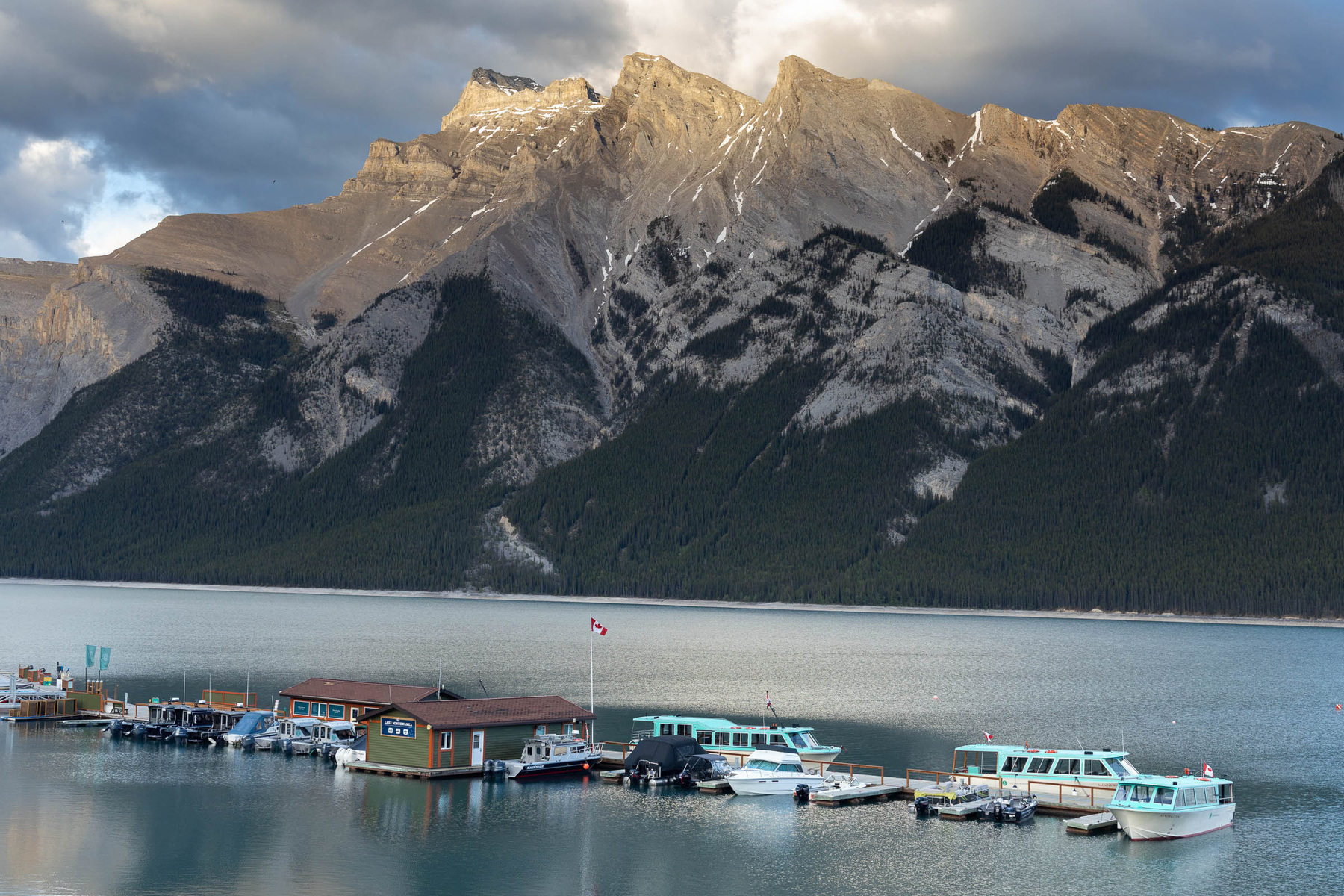 A serene mountain lake scene features boats docked at a pier with rocky mountains in the background.