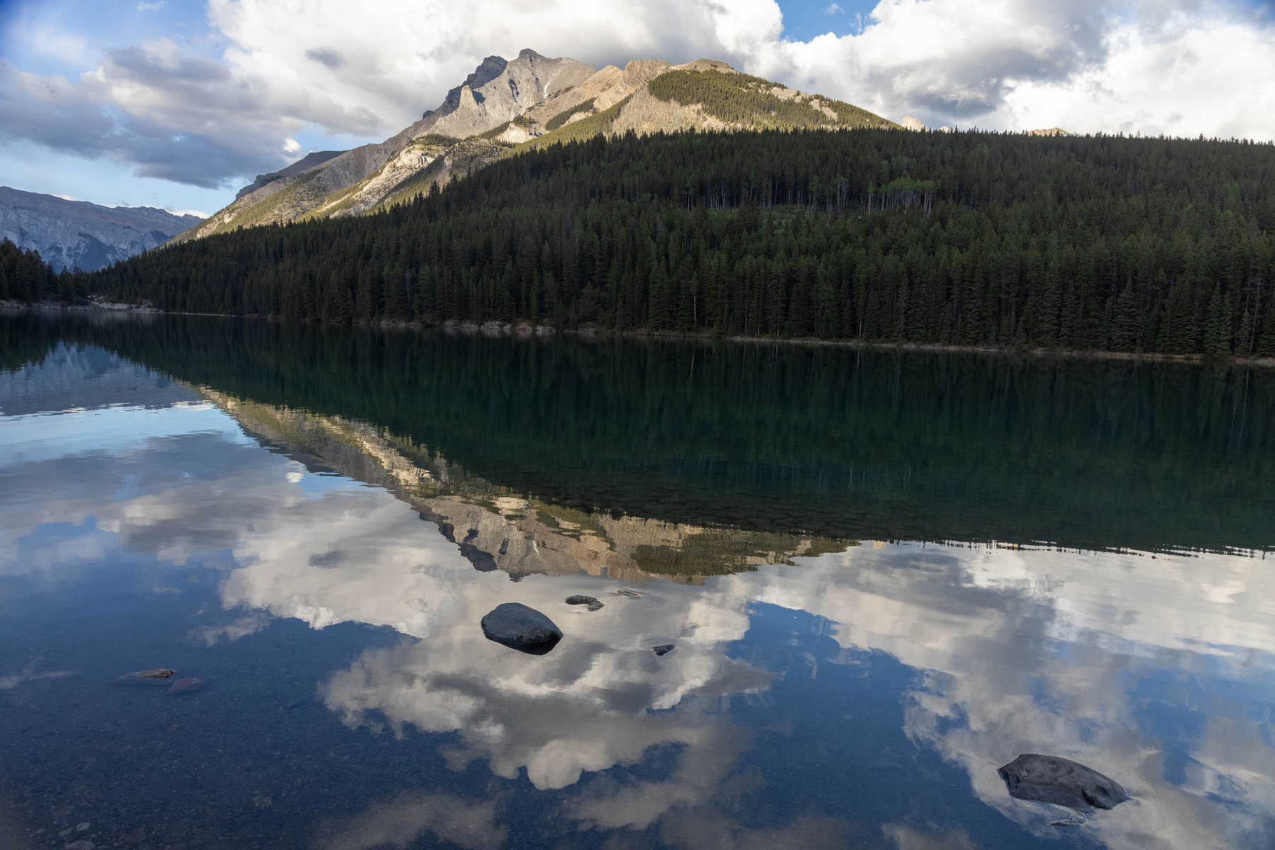 A serene mountain landscape is reflected in a calm lake, with a forested shoreline and scattered clouds in the blue sky.