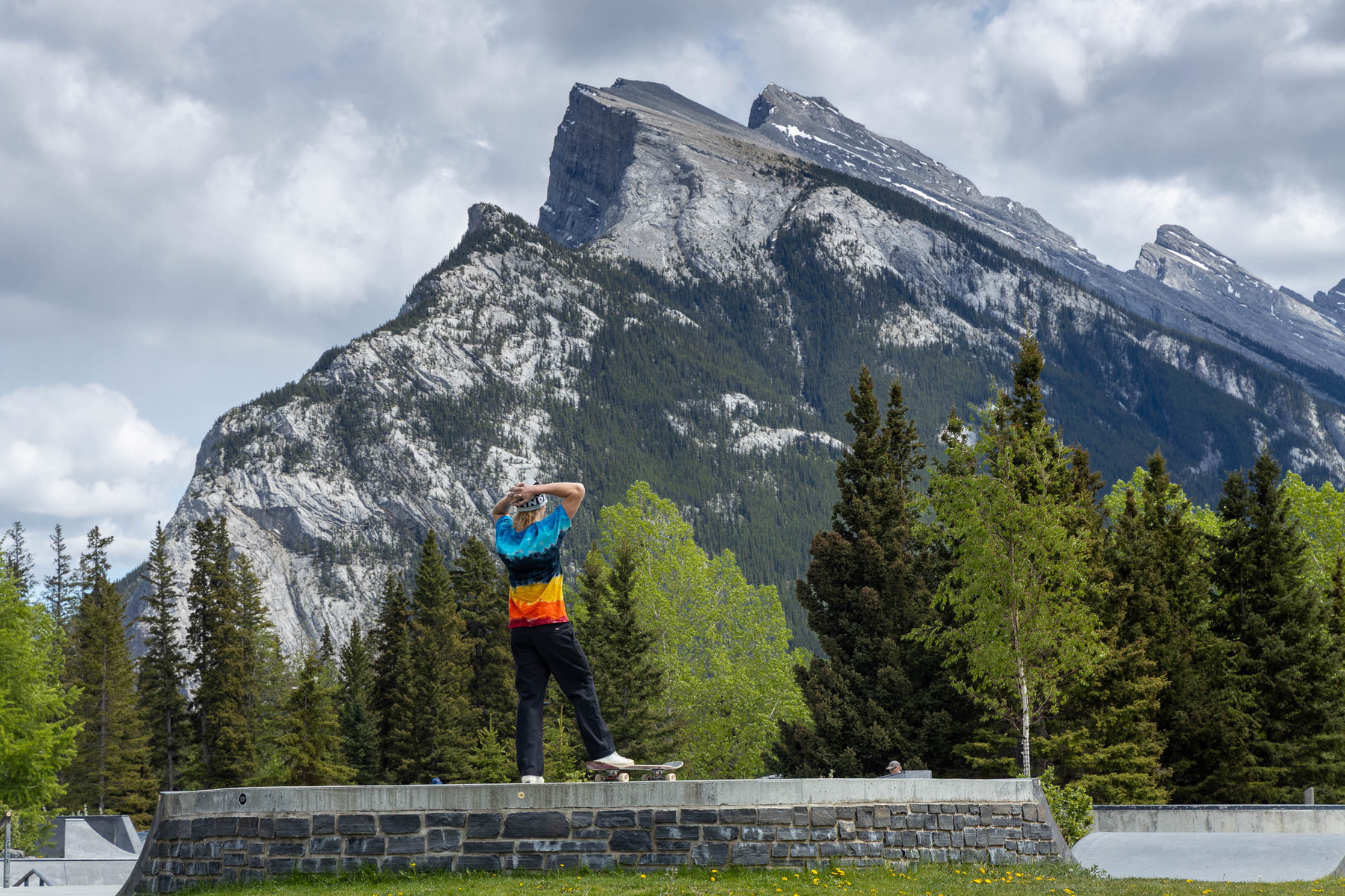 A person wearing a colorful shirt stands on a stone platform, gazing at a mountainous landscape with evergreen trees in the foreground.