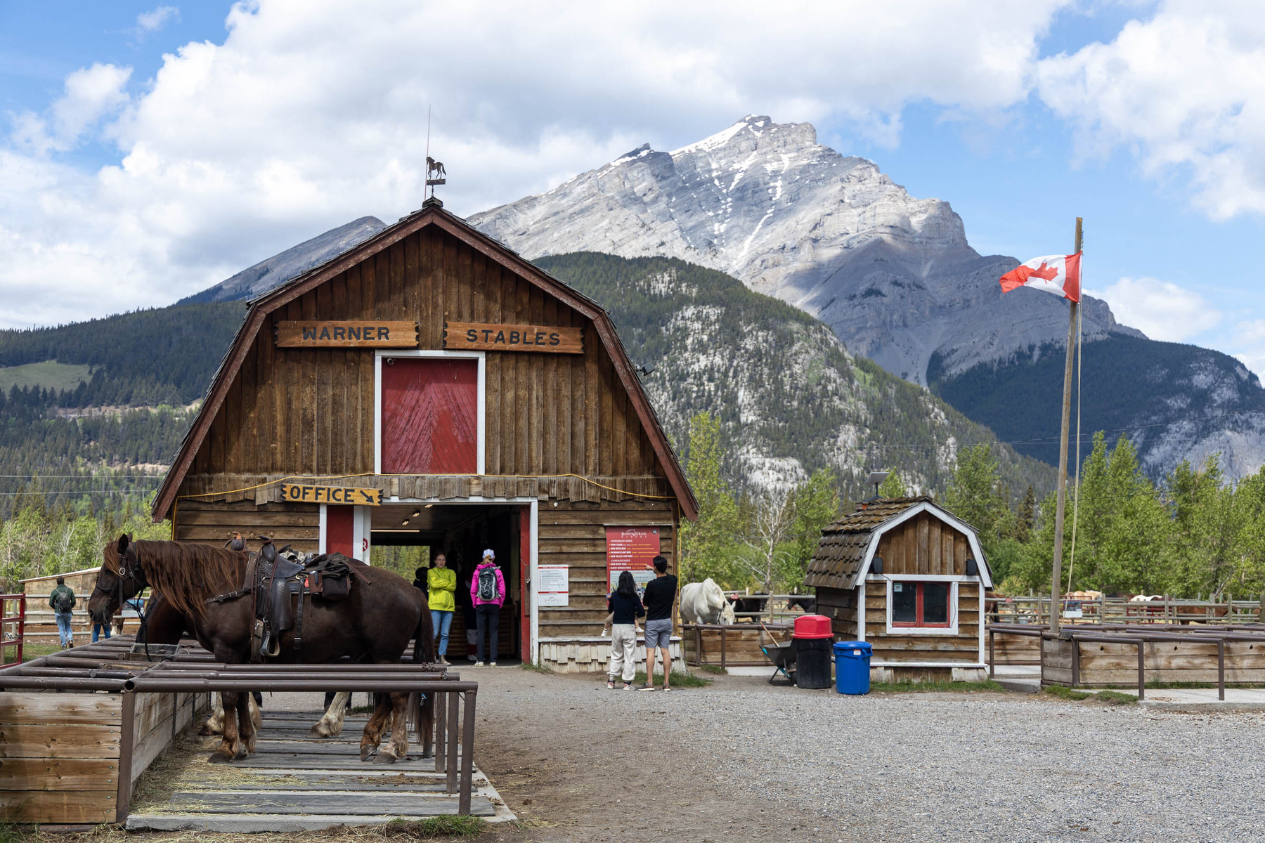 A rustic wooden stable with horses, people, and a Canadian flag is set against a backdrop of majestic mountains.