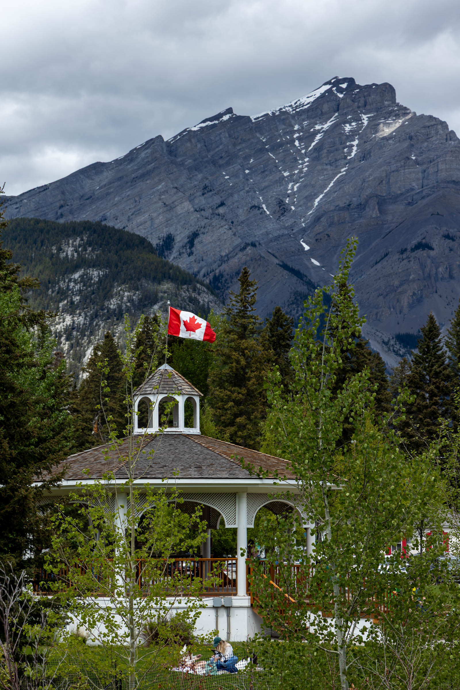 A gazebo with a Canadian flag sits amidst lush greenery, with snow-capped mountains in the background.